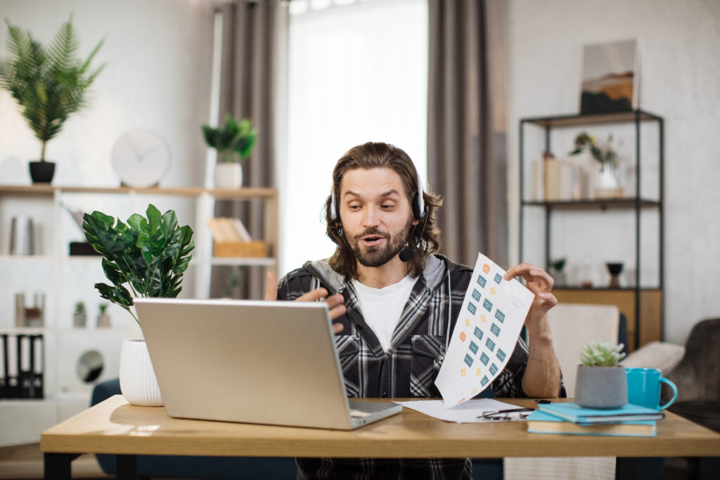 Creative male manager leading working meeting through video call on laptop. Young bearded man freelance showing graphs and chart while sitting at table in his office.